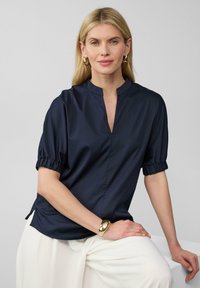 Tunic blu navy con un parziale scollo a bottone e maniche corte e plissettate. Tessuto liscio e leggero con una vestibilità rilassata, abbinata a pantaloni crema.