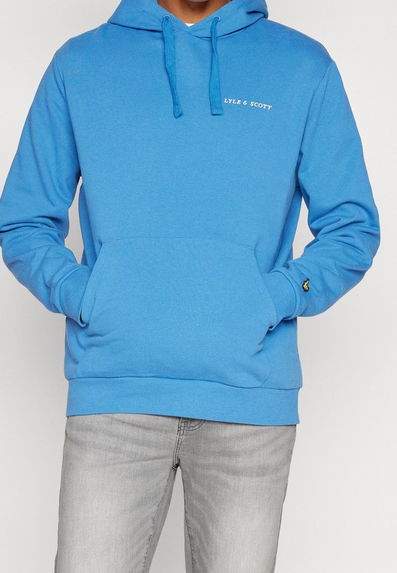 Felpa con cappuccio in cotone blu con tasca frontale, cappuccio regolabile con cordino e piccolo logo bianco sul petto. Abbinata a jeans grigio chiaro.