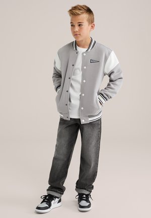 Un garçon se tenant debout avec les mains dans les poches, portant une veste varsity grise et blanche, un t-shirt blanc, un jean noir et des baskets noir et blanc.