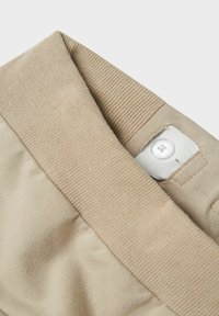 Pantalons beiges avec une taille côtelée, dotés d'une fermeture à bouton et de coutures plates pour une finition lisse. Le tissu semble doux et flexible.