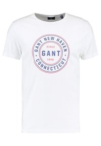 Camiseta de algodón blanca de manga corta, con un diseño gráfico circular en azul y rojo que incluye el texto "GANT NEW HAVEN CONNECTICUT" y la fecha.
