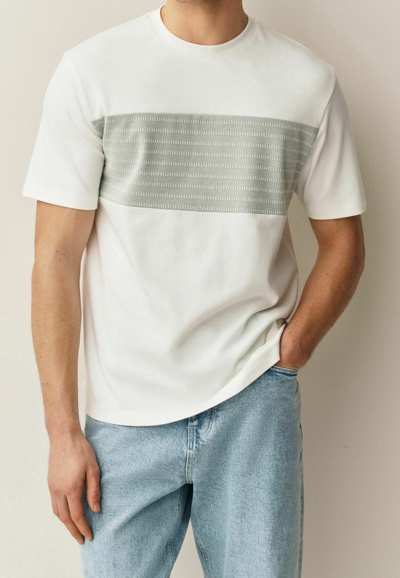 Homme portant une chemise blanche à manches courtes avec une large bande horizontale texturée gris clair sur la poitrine et un jean en denim bleu clair.