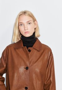 Brun læderjakke med en krave, store sorte knapper og en glat tekstur, lagdelt over en sort rullekrave.