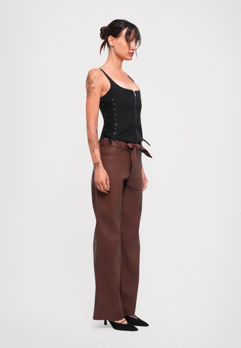 Vrouw met tatoeages draagt een zwarte rits-top met zijveters, bruin hoogwaist, wijde broek met een strikriem, en zwarte puntige hakken.