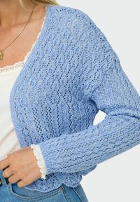 Cardigan a maglia blu chiaro con design aperto. Presenta un motivo in pizzo testurizzato e polsini a coste. Indossato sopra una maglietta bianca.