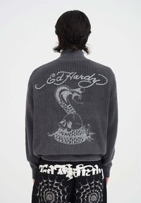 Mann in grau gestricktem Pullover mit weißem Totenkopf- und Schlangendesign sowie "Ed Hardy"-Aufschrift, kombiniert mit schwarzen Hosen mit Spinnennetzmustern.