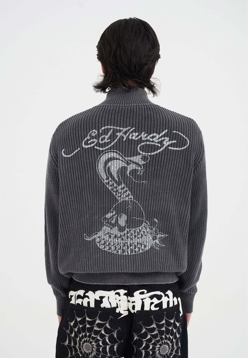 Mann in grau gestricktem Pullover mit weißem Totenkopf- und Schlangendesign sowie "Ed Hardy"-Aufschrift, kombiniert mit schwarzen Hosen mit Spinnennetzmustern.