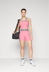 Rosa idretts crop top og shorts med grønne detaljer og svart Adidas-logo, kombinert med en svart treningsveske og hvite joggesko med striper.