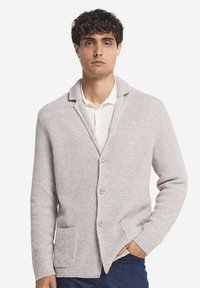 Jasnoszara kardigan z teksturowanym splotem, z wyciętym kołnierzem, pięcioma guzikami i kieszeniami z przodu, noszony na białej koszuli.