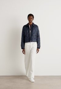 Marineblauwe gequiltte jas met een kraag, gecombineerd met een gestreept shirt, witte wijde broek en witte sneakers, op een neutrale achtergrond.