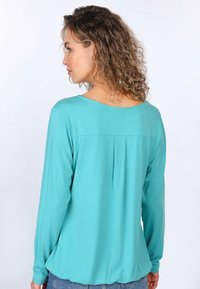 Türkises Langarmshirt mit lockerem Schnitt, rundem Ausschnitt und Falten-Detail auf der Rückseite. Weicher Stoff und leicht abgerundeter Saum.