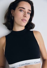 Soutien-gorge de sport noir à col montant en tissu lisse, avec une bande blanche portant le logo "Calvin Klein" en bas.