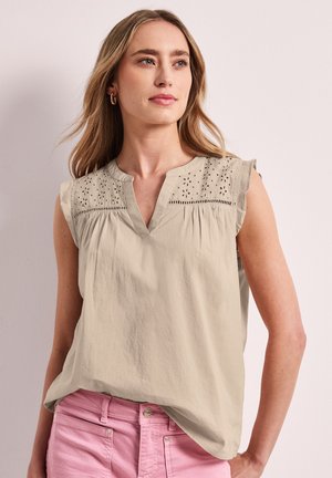 Femme aux longs cheveux blonds portant un chemisier beige sans manches avec des détails œillets et un pantalon rose, regardant légèrement vers la droite.