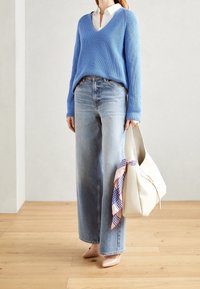 Woman wearing a blue V-neck sweater over a white shirt, light blue wide-leg jeans, beige heels, holding a cream bag with a patterned scarf.