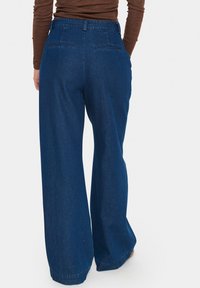 Platus džinsai tamsiai mėlynos spalvos, su aukštu juosmeniu ir dviem užpakalinėmis kišenėmis su subtiliu siuvinėjimu. Minkšta, tvirta medžiaga.