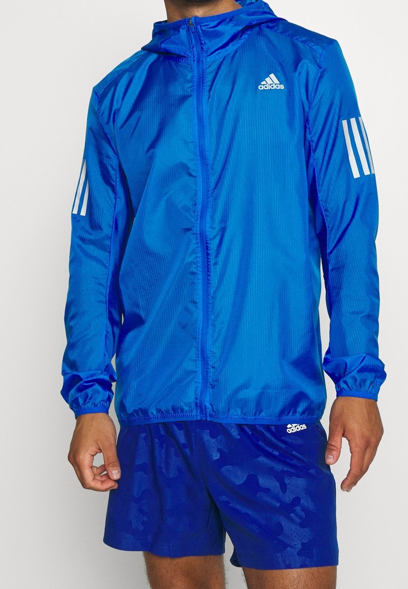 Mężczyzna w niebieskiej kurtce z kapturem Adidas z białymi paskami na rękawach oraz pasujących niebieskich sportowych spodenkach Adidas.