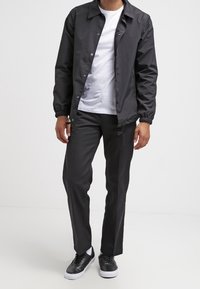 Svart lättviktsjacka med tryckknappar, vit T-shirt, svarta byxor med dragsko och svarta sneakers med vita sulor.