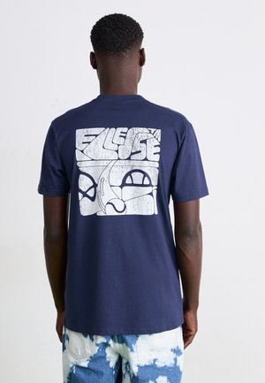 Mann trägt dunkelblaues T-Shirt mit großem weißem abstraktem Motiv und Text auf der Rückseite, kombiniert mit blau-weißen Tie-Dye-Shorts.