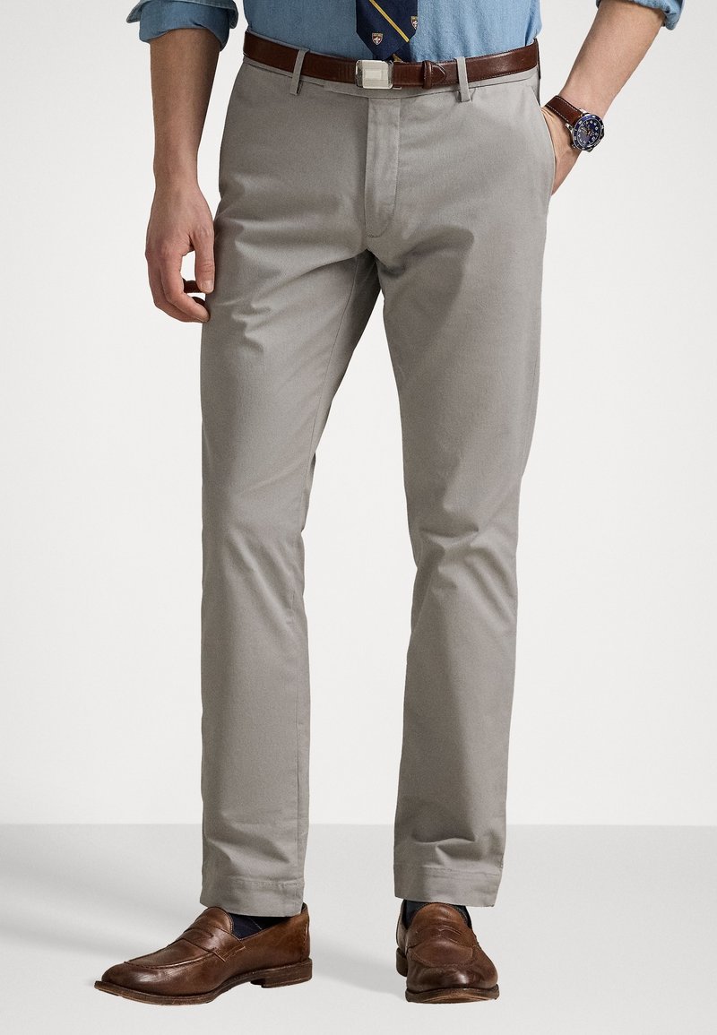Homme portant un pantalon gris clair avec une ceinture en cuir marron, des mocassins en cuir marron, une chemise bleue retroussée, une cravate à motif et une montre-bracelet.