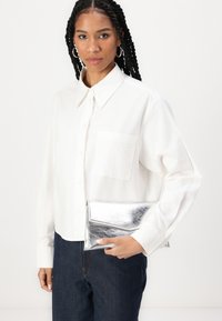 Camicia bianca a maniche lunghe con colletto e tasca, abbinata a una borsa a tracolla argentata e indossata con jeans scuri. Tessuto dalla texture liscia.