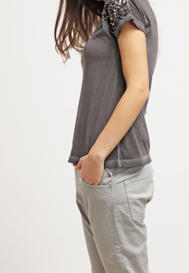 Femme portant un t-shirt gris avec des détails en sequins sur les manches et un pantalon gris clair, une main dans la poche, de profil.