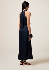Marineblaue Maxikleid mit Neckholder und gebundenem Rücken, weicher Textur, fließendem Schnitt, kombiniert mit schwarzen flachen Sandalen und einem Spiked-Cuff-Armband.