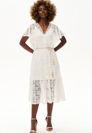 Kobieta o kręconych włosach, ubrana w białą koronkową sukienkę midi z falbanowymi rękawami, pasem ze sznurka i beżowe szpilki z paskiem wokół kostki, stoi z rękami na biodrach.