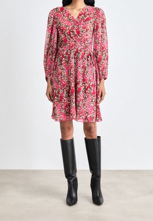 Robe portefeuille florale en tissu rose avec des manches longues ; présente une taille cintrée et une jupe à volants. Portée avec des bottes noires jusqu'au genou.