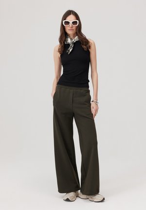 Kobieta w białych okularach przeciwsłonecznych, czarnym topie bez rękawów, kraciastym szaliku na szyi, oliwkowych szerokich spodniach i beżowych sportowych butach, stojąca z rękami w kieszeniach.