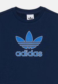 Sötétkék, rövid ujjú pamut póló. Kék Adidas logóval rendelkezik, három levélre emlékeztető akcentussal és vízszintes csíkokkal alatta.