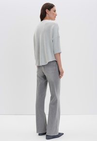 Frau steht seitlich mit leicht grauem strukturiertem Pullover, ausgebleichten grauen geraden Jeans und dunkelblauen Schuhen vor einem einfarbigen Hintergrund.