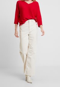 Pull rouge à col en V et pantalon large en velours côtelé crème à rayures verticales ; le modèle porte des chaussures à talons beiges. Design simple, coupe décontractée.