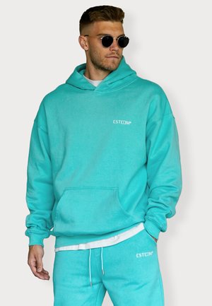 Helle türkisfarbene Kapuzenjacke aus weichem Material, mit einer Fronttasche und Kordelzug-Haube. Weißes Logo auf der Brust. Kombiniert mit passenden Hosen.