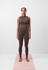 Smešano smeđi uski crop top i hlače visokog struka izrađene od rastezljivog materijala. Model stoji bos na ružičastom yogamat-u nasuprot jednobojnoj pozadini.