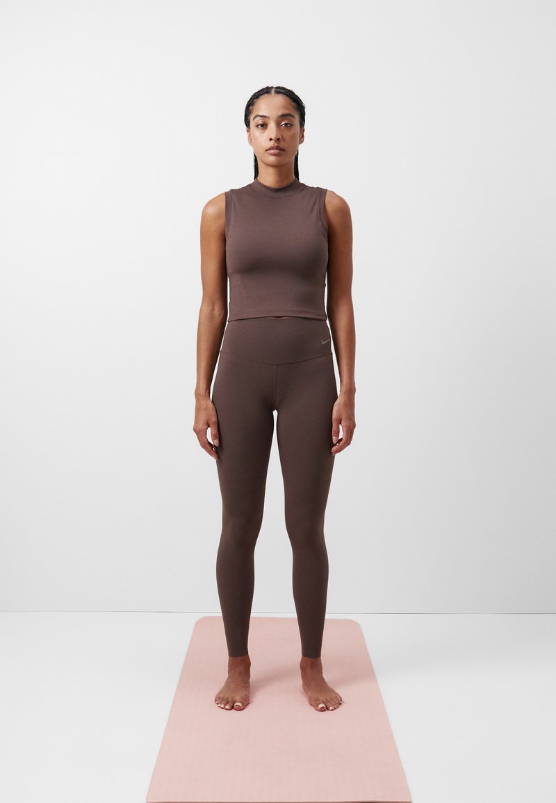 Smešano smeđi uski crop top i hlače visokog struka izrađene od rastezljivog materijala. Model stoji bos na ružičastom yogamat-u nasuprot jednobojnoj pozadini.