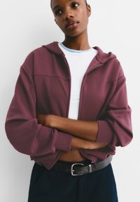 Weinroter Reißverschluss-Hoodie mit lockerem Schnitt, gerippten Bündchen und abgerundetem Saum, über einem weißen Hemd getragen. Dunkle Hose und ein schwarzer Gürtel vervollständigen den Look.