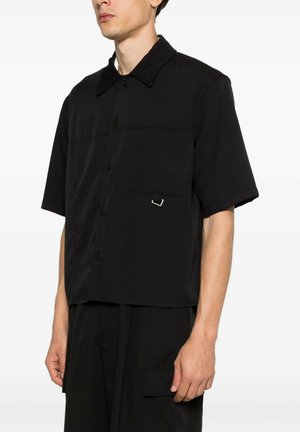 Uomo che indossa una camicia nera morbida a maniche corte con bottoni e taschino sul petto con un piccolo dettaglio in fibbia argento.