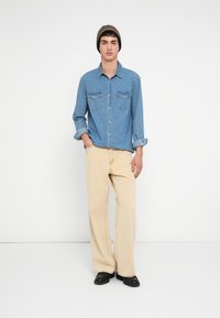 Jasnoniebieska koszula jeansowa z dwoma kieszeniami na piersi, rękawy podwinięte; w połączeniu z luźnymi beżowymi spodniami i czarnymi butami, model nosi brązową dzianą czapkę.