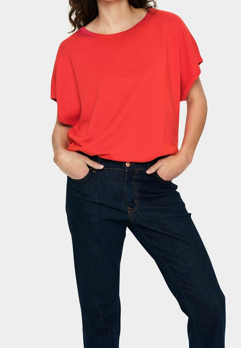 T-shirt rouge à manches courtes avec un col rond, associée à un jean en denim foncé avec des coutures orange et des poches latérales.