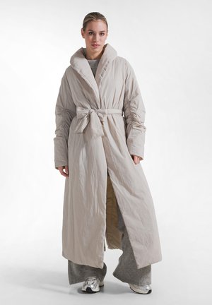Donna che indossa un lungo cappotto beige con cintura e maniche arricciate sopra pantaloni grigi larghi e sneakers argento, in piedi su sfondo bianco.