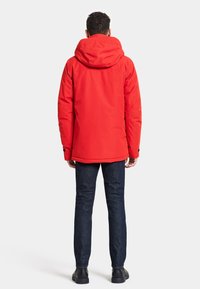 Röd isolerad jacka med huva, med en slät, vattentålig yta. I kombination med mörk denimjeans och svarta skor.