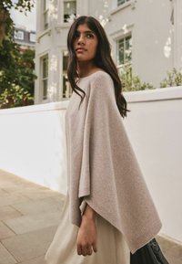 Lysegrå strikket poncho med brede ærmer, der har en blød tekstur. Kombineret med en flydende beige kjole, på en belagt gangsti nær hvide vægge.