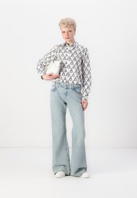 Hvit skjorte med svart geometrisk mønster, høyt liv lyseblå vide jeans og en liten hvit håndveske. Modellen har på seg hvite joggesko.