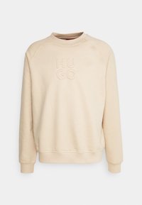 Sudadera beige con cuello redondo y mangas raglán. Presenta un logotipo "HUGO" en relieve en la parte delantera. Textura suave, ajuste casual.