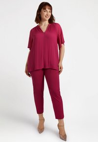 Fuchsias Top mit plissiertem Ausschnitt, kurzen Ärmeln und lockerer Passform; kombiniert mit fuchsia-farbenen Slim-Fit-Hosen; das Model trägt transparente Absatzschuhe.