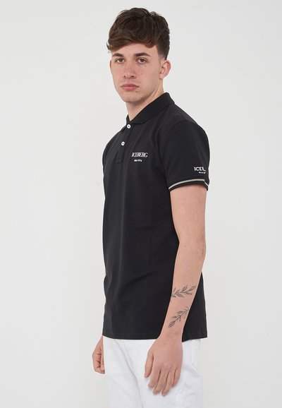 Polo shirt nera in cotone, con colletto, maniche corte con bordatura a contrasto e un logo sul petto. Sfondo bianco.