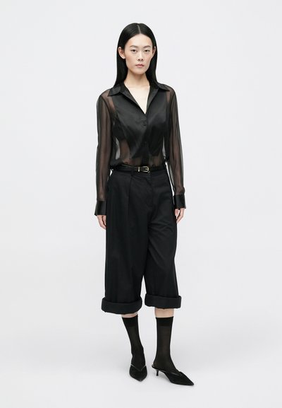 Black sheer button-up shirt worn with high-waisted black trousers, featuring rolled cuffs. Black heels and sheer black socks complete the outfit.