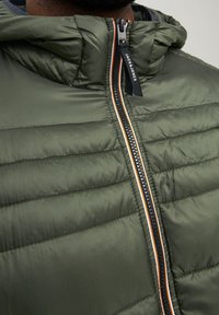 Chaqueta acolchada de color verde oliva con cierre de cremallera, capucha y rayas de acento en contraste en naranja y blanco a lo largo de la cremallera. Tejido texturizado.