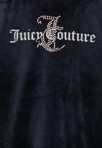 Črn žametni material z napisom "Juicy Couture" v velikih srebrnih kristalih, ki prikazuje dekorativno, pisan črko "J."