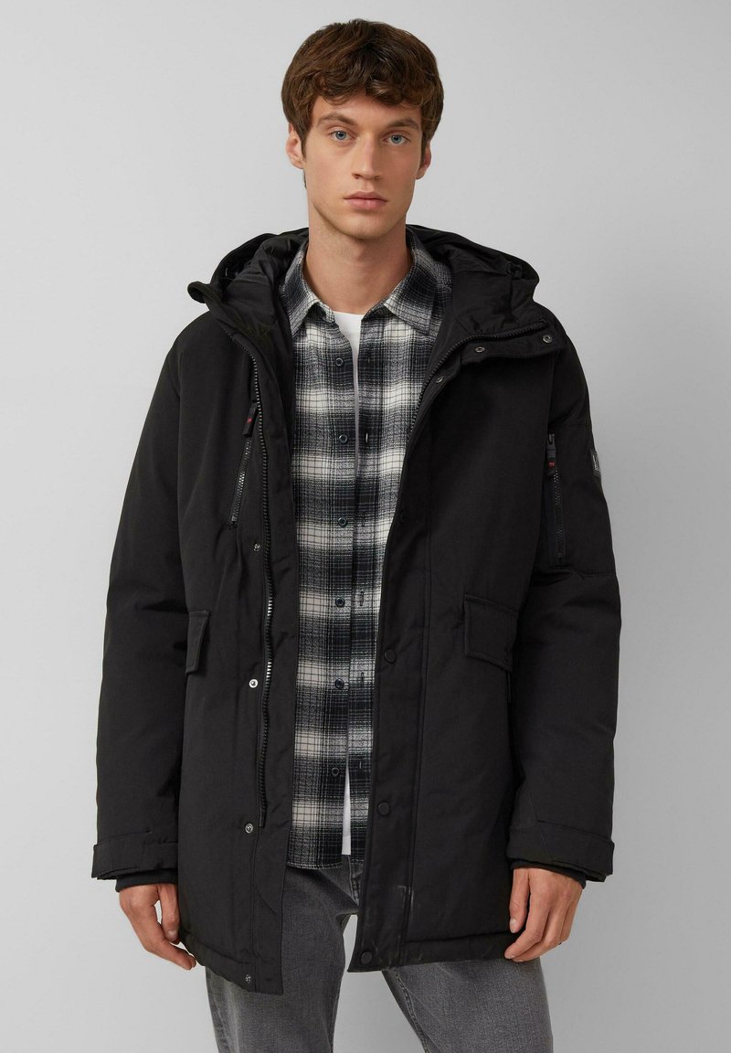 Schwarze gepolsterte Parka mit Kapuze, ausgestattet mit mehreren Taschen und einem Reißverschluss. Über einem grau und schwarz karierten Hemd getragen. Glatte Textur.